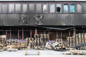 Exterior of a burned-out building with charred signage and stacks of wooden pallets in front, reflecting damage and aftermath of a fire.