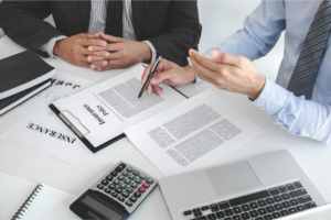 Two professionals reviewing home and auto insurance coverage documents at a desk with a calculator, laptop, and paperwork.